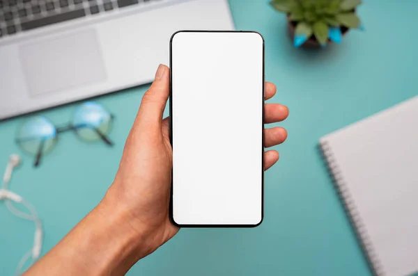 手に空白の画面を持つスマートフォンのクローズアップ 背景にはノートパソコン メモ帳があります デジタルガジェットを使う少女 学生の勉強やオフィスワーク 自宅からの仕事 フリーランスのための概念 トップビュー — ストック写真