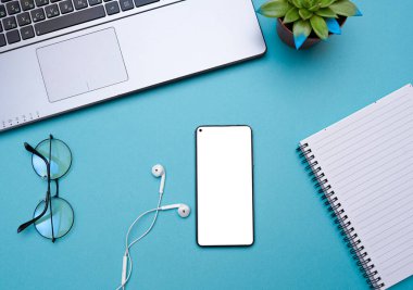 Beyaz ekranlı, kulaklıklı, laptoplu, mavi masadaki bloknotlu akıllı telefon. Çalışırken ya da ders çalışırken müzik dinleme kavramı. Yukarıdan bak. kopyalama alanı. Tasarım için model 