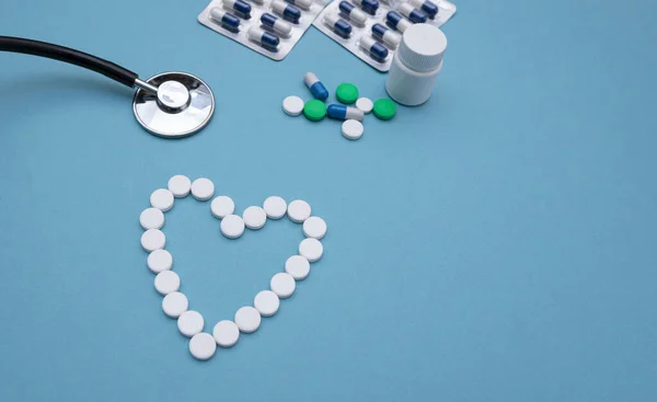 心形的白丸靠近听诊器 背景为蓝色背景特写 背景为多种药物 心脏病治疗的概念 药物与健康 把你的文字放好 顶部视图 — 图库照片