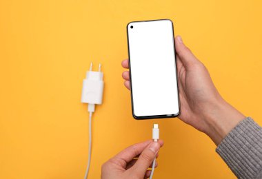 Akıllı telefonu USB kablosuyla şarj etmek ve arka planda sarı bir elle güç sağlamak şarj cihazını bağlar. Beyaz ekranlı akıllı telefon. Üst görünüm, iş ve teknoloji.