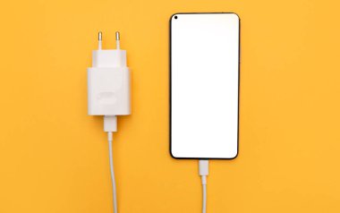 Smartphone sarı arka planda USB kablosu ve güç kaynağı ile şarj ediliyor. Beyaz ekranlı akıllı telefon. Üstten görüş, iş ve teknoloji. kopyalama alanı. Düz yatıyordu. Düzen