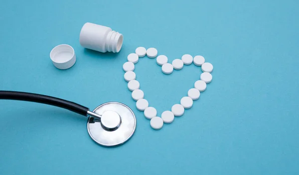 心脏形状的白色药丸从听诊器附近的罐子里倒出来 蓝色背景特写 心脏病治疗的概念 平躺在床上药物与健康 复制空间 顶视图 选择性重点 — 图库照片