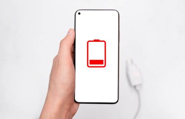 Akıllı telefon ellerinde ve beyaz ekranda boşaltılmış bir telefon bataryasının bir simgesi, arka planda bir blok ve pili şarj etmek için bir kablo. Seçici odaklanma. Üst Manzara. metin için boşluk kopyala. maketleme