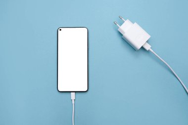 Mavi arka planda USB kablosu ve güç kaynağı üzerinden akıllı telefon şarj ediliyor. Beyaz ekranlı akıllı telefon. Kablolu şarj edilebilir telefon kavramı. kopyalama alanı. Düz yatıyordu. maketleme