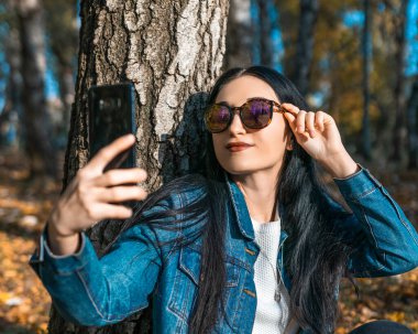Güneş gözlüklü mutlu genç kadın, sonbaharda ya da kış parkında bir ağacın yanında cep telefonu kullanarak selfie çekiyor. Sonbaharda parkta telefon kullanan esmer kız.. 