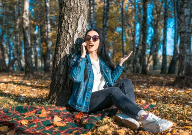 Bir esmer telefonda duygusal iletişim kuruyor, güneş gözlüklü şaşırmış bir kadın sonbaharda bir ağacın yanındaki parkta dinleniyor. Doğada akıllı telefondan iletişim kavramı.