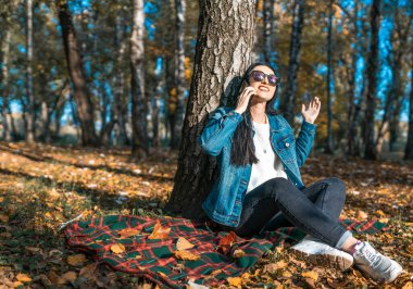 Esmer bir kadın cep telefonuyla duygusal olarak iletişim kuruyor, güneş gözlüklü şaşırmış bir kadın sonbaharda bir ağacın yanındaki parkta dinleniyor. Doğadaki akıllı telefondan iletişim konsepti. Boşluğu kopyala