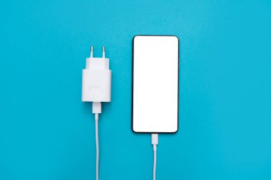 Beyaz telefon ekranı ve güç kaynağı olan akıllı telefon, mavi arka planda şarj aleti. Telefonun bataryasını şarj etmek için USB kablosu. Düz konum, üst görünüm, metin için boşluk kopyala