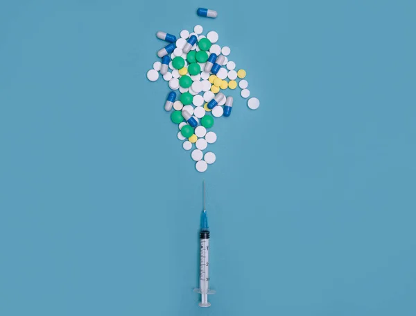 Mavi arka planda iğne ve haplarla dolu bir şırınga. Şırınga iğnesinden çeşitli tabletler ve kapsüller çıkıyor. Vitamin kompleksi konsepti, bir çok hapın yerine bir şırınga antibiyotikle değiştirilmesi.