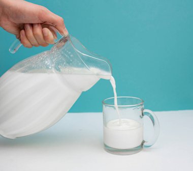 Fresh cow's milk is poured from a jug into a mug on a white and blue background. Milk Day. Healthy and tasty breakfast concept. copy space for text