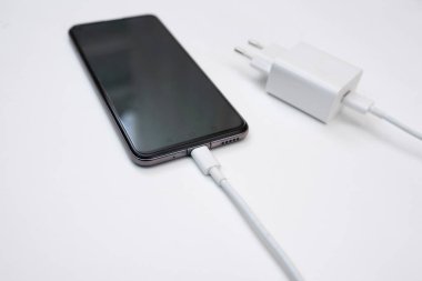 Beyaz arka planda USB kablosuyla akıllı telefonu şarj etmek, yakın plan. Beyaz arka planda USB şarj kablosu ile asılı duran siyah bir akıllı telefonun silueti. Boşluğu kopyala
