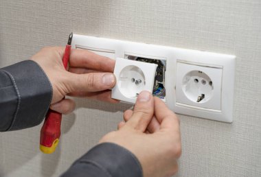 Üniformalı elektrikçi odadaki beyaz duvardaki elektrik prizlerini topluyor. Bir elektrikçi daireye tornavida kullanarak soket takıyor. Dairede tadilat var..