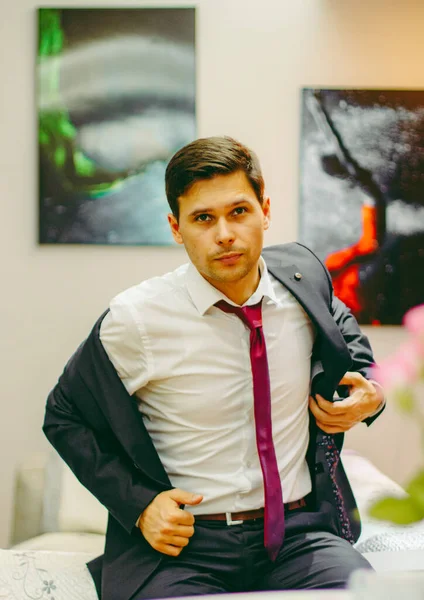 Yatağın kenarında oturan takım elbiseli ve kırmızı kravatlı adam. Takım elbiseli adam odada. Siyah takım elbiseli ve kırmızı kravatlı adam. Adam ceketini çıkardı.