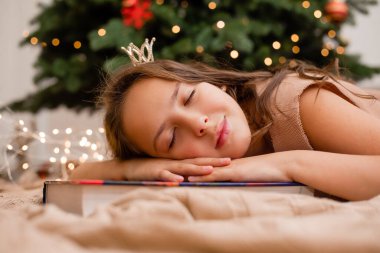 Prenses kostümü giymiş güzel bir kız masallarla dolu bir kitaptaki Noel ağacının yanında uyuyakalmış.