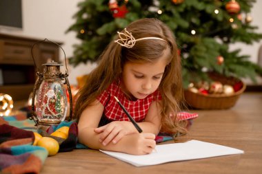 Kırmızı elbiseli şirin küçük kız Noel Baba 'ya mektup yazıyor evde Noel ağacının yanında.