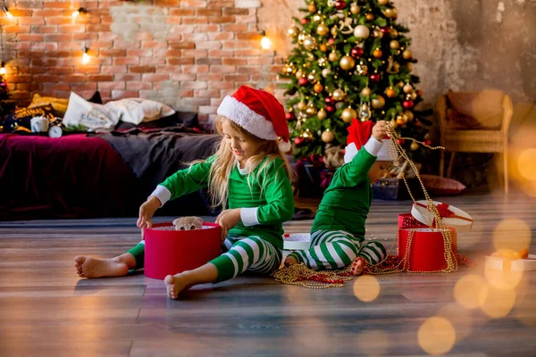 两个身穿睡衣 头戴圣诞帽的小孩在房间里打开了圣诞礼物的包装 — 图库照片