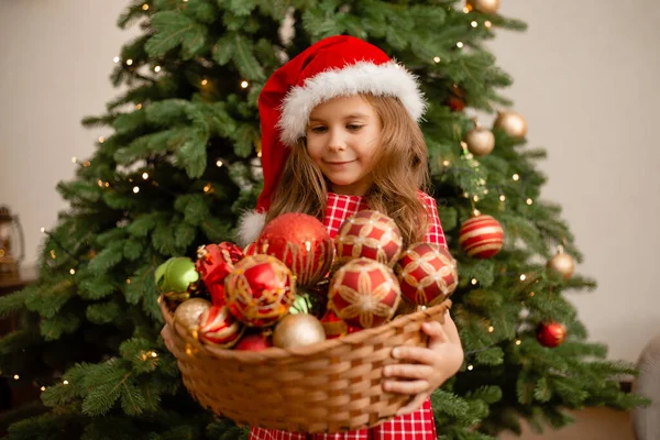 在圣诞树下 戴着圣诞礼帽的可爱小女孩拿着一篮子玩具 — 图库照片