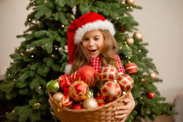 在圣诞树下 戴着圣诞礼帽的可爱小女孩拿着一篮子玩具 — 图库照片