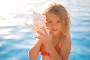 Küçük kız akşam güneşinde havuz kenarında büyük deniz kabuğuyla poz veriyor.