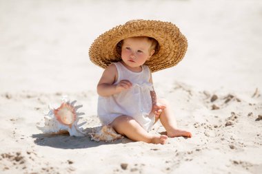 Hasır şapkalı şirin kız deniz kabuklarıyla kumların üzerinde oturuyor. 