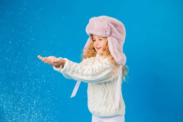 cute little girl in pink furry hat playing with fake snow on blue background in studio