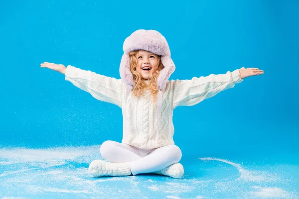 cute little girl in pink furry hat playing with fake snow on blue background in studio