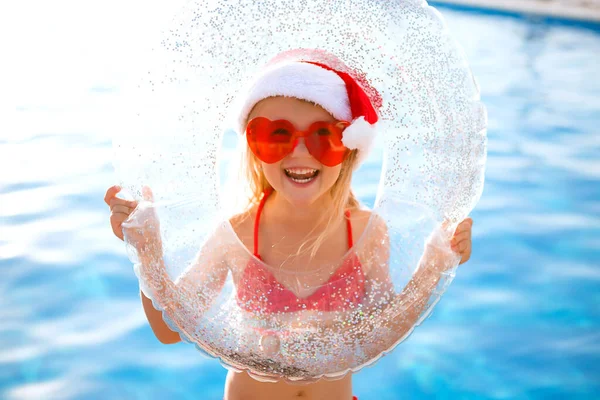 Gülümseyen, kalp şeklinde gözlüklü küçük kız ve havuz kenarında yüzme çemberi tutan Noel şapkası.