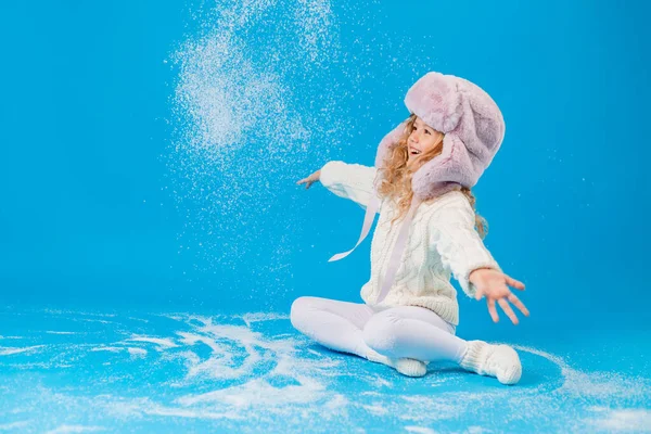 cute little girl in pink furry hat playing with fake snow on blue background in studio