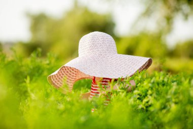 Küçük kız büyük bir şapka takıyor. Yeşillik içinde saklanıyor.