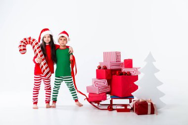 Noel kostümlü tatlı kız ve oğlan şeker kamışı balonuyla poz veriyorlar ve kızakta bir sürü hediye var.