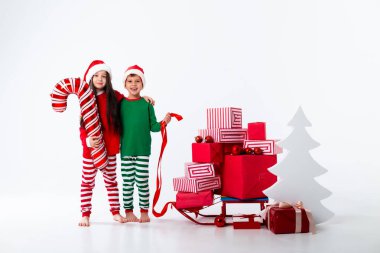 Noel kostümlü tatlı kız ve oğlan şeker kamışı balonuyla poz veriyorlar ve kızakta bir sürü hediye var.