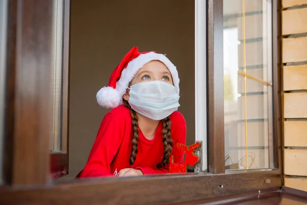 Tıbbi maskeli ve Noel Baba şapkalı kız pencereden dışarı bakıyor.
