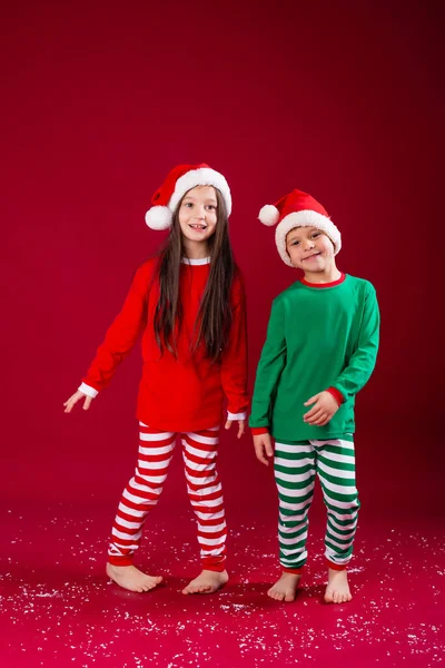 两个穿着节日服装戴着圣诞礼帽的可爱孩子在红工作室的背景下跳舞 — 图库照片