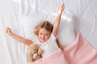 Küçük kız pembe ekose ve oyuncak ayıyla yatakta.