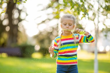 Çok renkli bir bluz giymiş, yaz parkında sabun köpüğü üfleyen tatlı küçük bir kız.