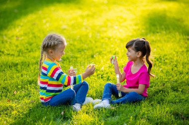 İki küçük kız yaz çimlerinde oturup sabun köpüğü üflüyor. Avrupa ve Hindistan etnik kökenli çocuklar