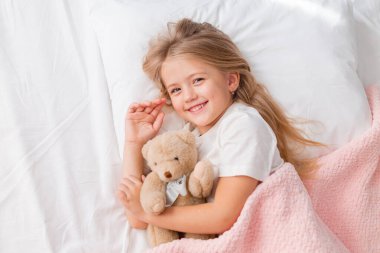 Küçük kız pembe ekose ve oyuncak ayıyla yatakta.