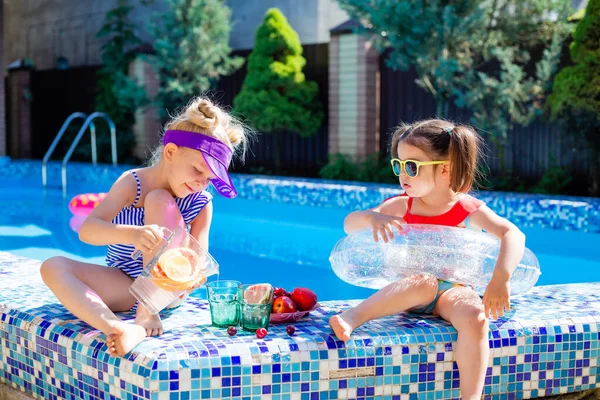 Cute girls sitting close to pool, having fun, eating fruits and drinking lemonade. Summer vacation concept