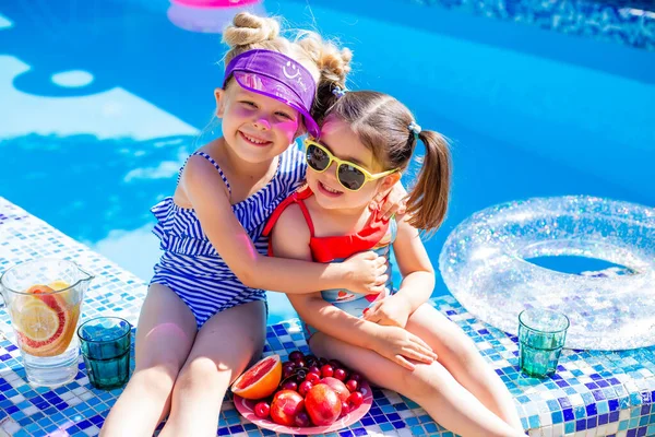 Cute girls sitting close to pool, having fun, eating fruits and drinking lemonade. Summer vacation concept