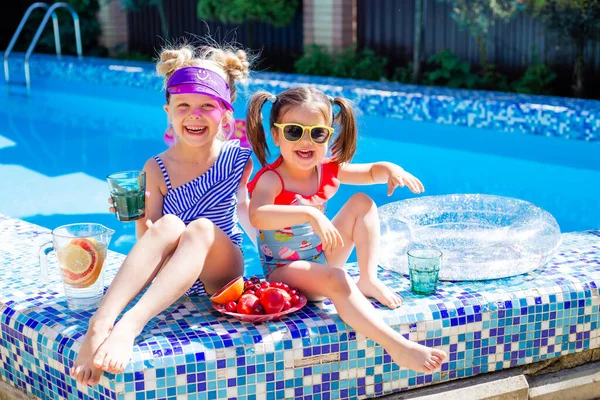 Cute girls sitting close to pool, having fun, eating fruits and drinking lemonade. Summer vacation concept
