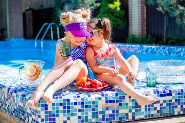 Cute girls sitting close to pool, having fun, eating fruits and drinking lemonade. Summer vacation concept