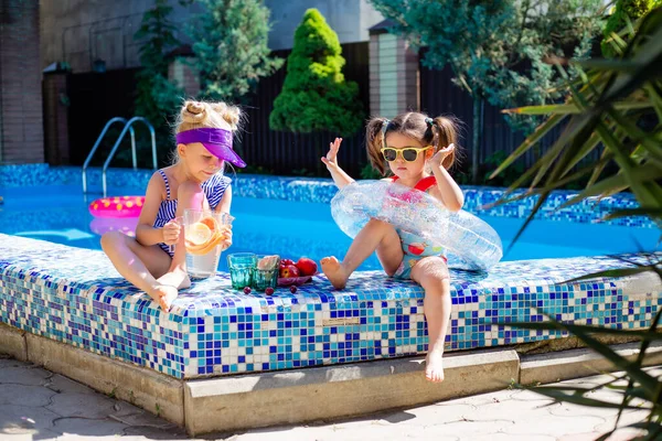 Cute girls sitting close to pool, having fun, eating fruits and drinking lemonade. Summer vacation concept