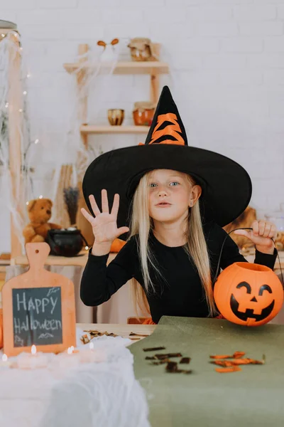 彼女の手にカボチャの形のキャンディーバスケットを保持している巨大な魔女の帽子と魔女の衣装で長い髪を持つ小さなブロンドの女の子の肖像画 ハロウィンのコンセプト テキスト用のスペース 高品質の写真 — ストック写真