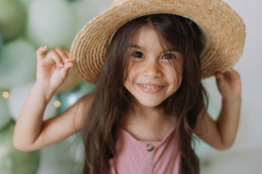 Pembe elbiseli şirin bir kız çocuğu ve hasır şapkalı doğum gününü kutlayan renkli balonlar.