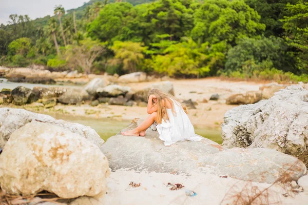 Chica triste ofendido con el pelo largo sentado en una roca en la orilla del mar hacia atrás y pensando en algo — Foto de Stock