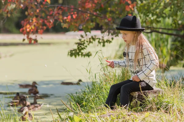 Mädchen mit Hut sitzt auf einem Baumstumpf in einem Park sonnigen Sommertag, Blick auf den See, wo Enten — Stockfoto