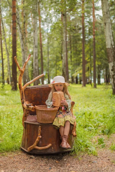 Mädchen in Hut und Kleid rustikal und Stiefel sitzen auf einem Baumstumpf im Wald mit einem Korb mit Brot und Brötchen — Stockfoto