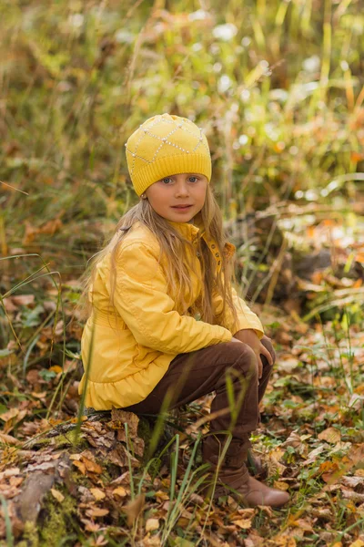 Menina em uma jaqueta amarela e um chapéu amarelo sentado em um toco de árvore na floresta de outono um dia ensolarado — Fotografia de Stock