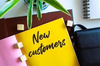Ofis masasında ofis malzemeleriyle birlikte NEW CUSTOMERS yazan defter. Sarı renk arkaplanı.