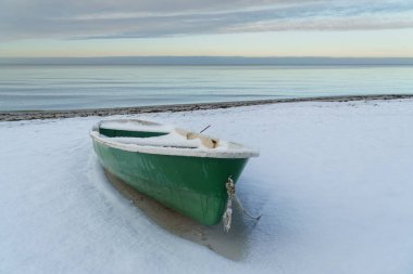 A quiet winter scene featuring an abandoned  fishing boat resting on a snow-covered sandy beach with a small bare tree, framed by a calm sea and forest horizon